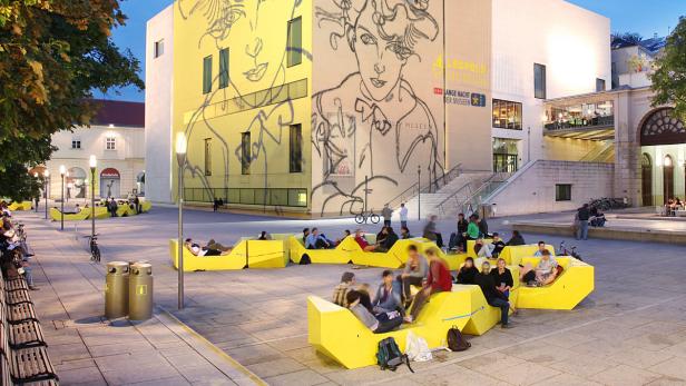 Menschen sitzen auf gelben Sitzgelegenheiten vor dem Leopold Museum in Wien.