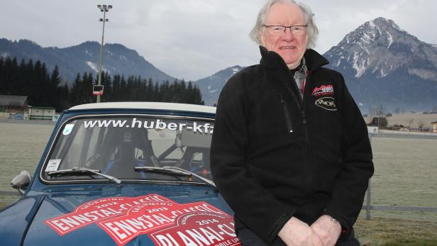 Ein Mann steht vor einem blauen Oldtimer mit Aufklebern der Ennstal-Classic Rallye vor einer Bergkulisse.