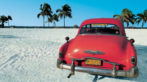 Ein roter Oldtimer von Chevrolet steht an einem Strand mit Palmen im Hintergrund.