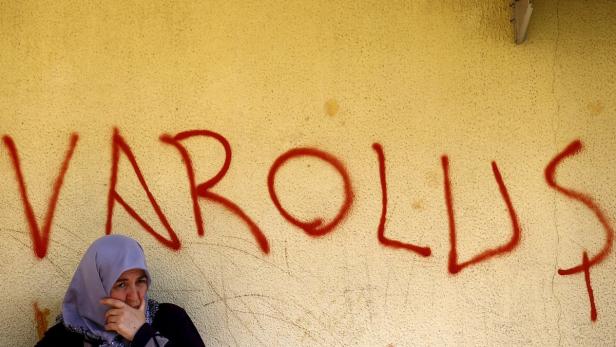 Eine Frau mit Kopftuch steht vor einer Wand mit dem Graffiti „VAROLUŞ“.
