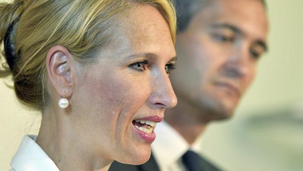 Eine blonde Frau mit einem Perlenohrring, im Hintergrund ein Mann.