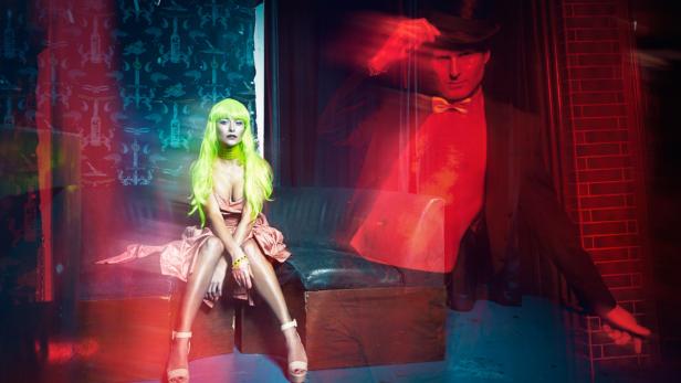 Eine Frau mit grüner Perücke sitzt vor dem transparenten Abbild eines Mannes im Anzug.