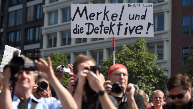 Eine Menschenmenge mit Kameras und einem Schild mit der Aufschrift „Merkel und die Detektive!“.