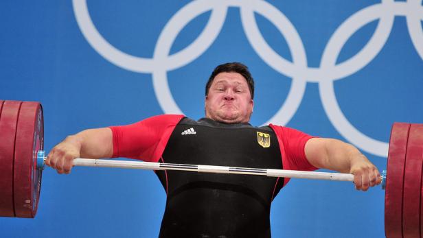 Ein Gewichtheber stemmt eine schwere Hantel vor dem Hintergrund der olympischen Ringe.