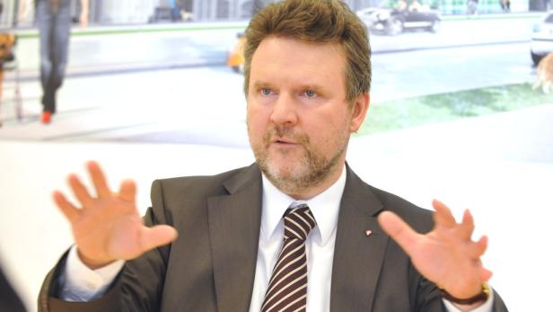 Ein Mann im Anzug gestikuliert vor einem unscharfen Hintergrund mit einer Straßenszene.
