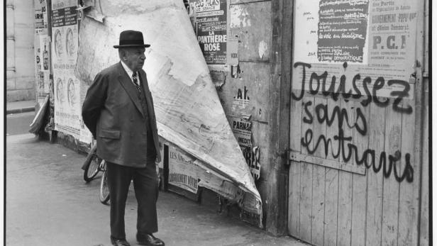 Ein Mann mit Hut steht vor einer Wand voller Plakate und Graffiti in Paris.