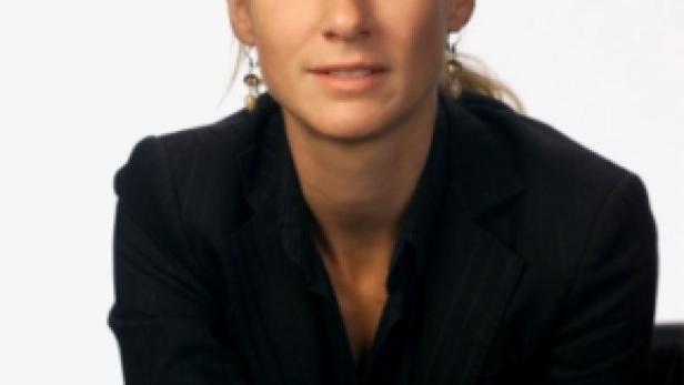 Eine Frau mit blonden Haaren und dunklem Blazer sitzt auf einem Stuhl.