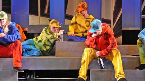 Eine Gruppe junger Schauspieler sitzt in farbenfrohen Kostümen auf einer Bühne.