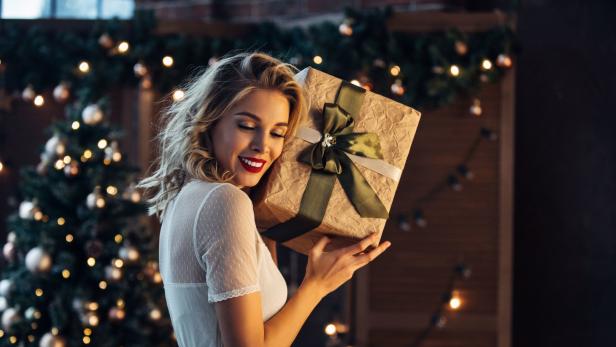 Eine Frau hält freudig ein Geschenk vor einem festlich geschmückten Weihnachtsbaum.