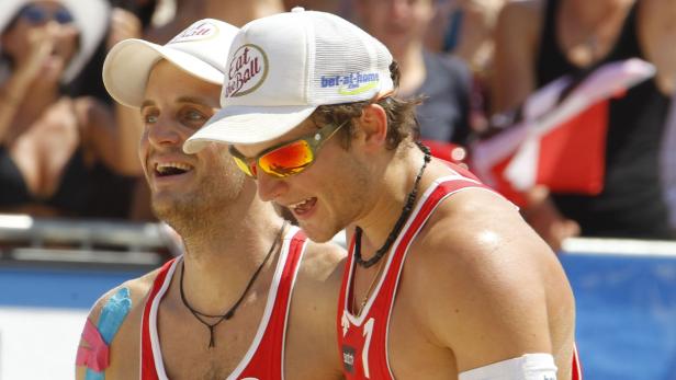 Zwei Beachvolleyballspieler mit Kappen und Sonnenbrillen lächeln.