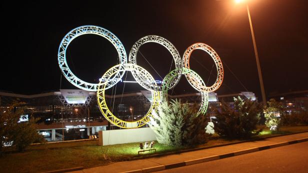 Die beleuchteten olympischen Ringe stehen vor einem Gebäude bei Nacht.