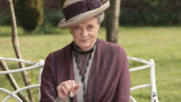 Maggie Smith als Violet Crawley in „Downton Abbey“ sitzt auf einer Bank.
