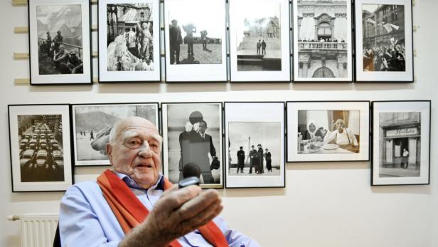 Ein älterer Mann sitzt vor einer Wand mit gerahmten Schwarzweißfotografien.