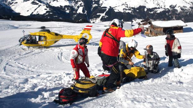 Ein gelber Rettungshubschrauber steht auf einer schneebedeckten Bergwiese, während Sanitäter eine Person versorgen.