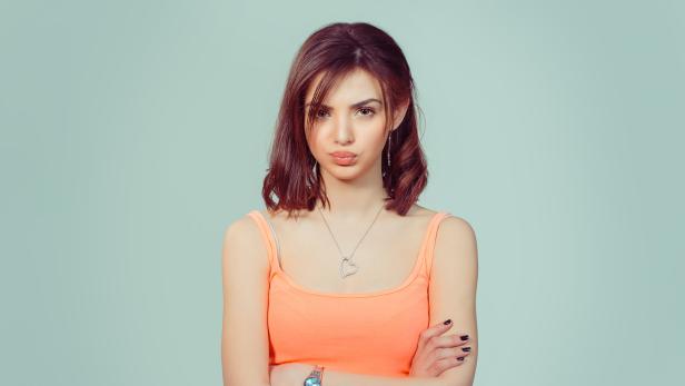 Eine Frau mit verschränkten Armen und einem schmollenden Gesichtsausdruck blickt in die Kamera.