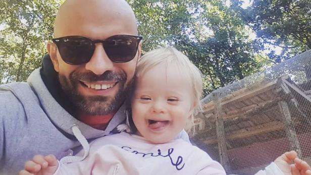 Ein lächelnder Mann mit Sonnenbrille posiert mit einem kleinen Mädchen mit Down-Syndrom vor einem Gehege.