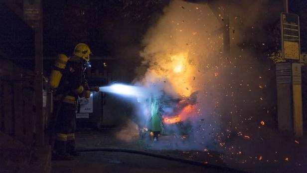 Ein Feuerwehrmann löscht einen brennenden Müllcontainer bei Nacht.