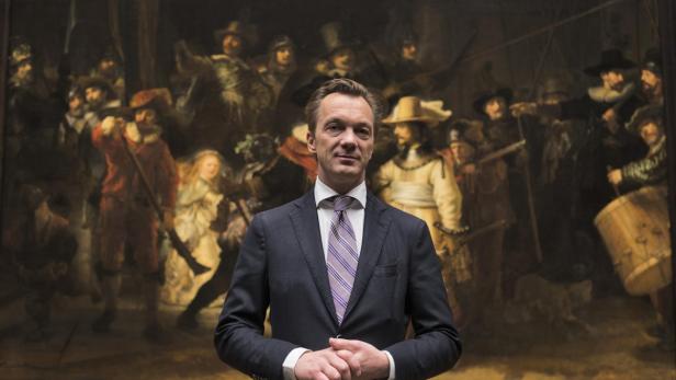 Ein Mann in Anzug steht vor Rembrandts Gemälde „Die Nachtwache“.