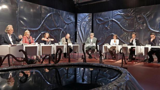 Acht Personen sitzen an Tischen vor einer silbernen Wand während einer Fernsehdiskussion.