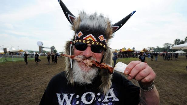 Ein Mann mit Fellmütze und Sonnenbrille isst einen Fleischspieß auf dem Wacken Open Air Gelände.