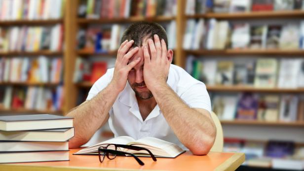 Ein Mann sitzt verzweifelt mit einem Buch und einer Brille an einem Tisch in einer Bibliothek.