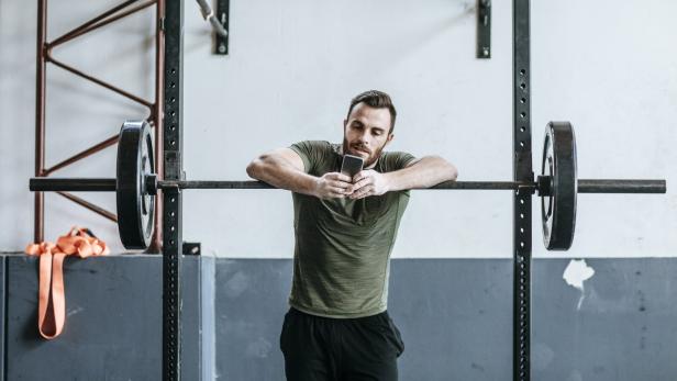 Ein Mann lehnt sich an eine Langhantelstange im Fitnessstudio und schaut auf sein Smartphone.
