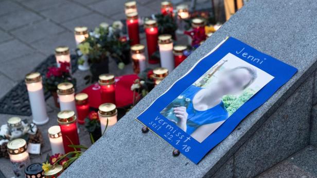 Ein Suchplakat für die vermisste Jenni liegt auf einer Steinmauer, umgeben von zahlreichen brennenden Grablichtern und Blumen.