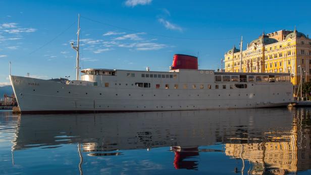Die Yacht „Marina“ liegt vor einem gelben Gebäude im Hafen.
