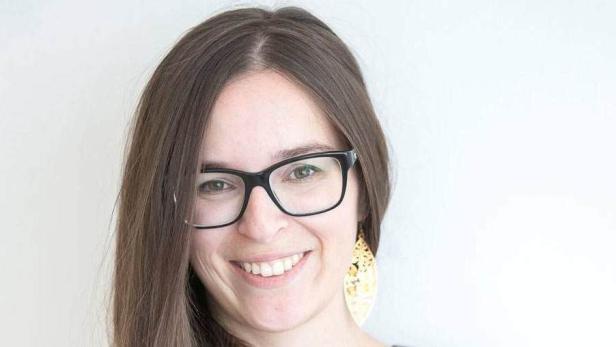 Eine lächelnde Frau mit Brille und goldenen Ohrringen vor einem hellen Hintergrund.
