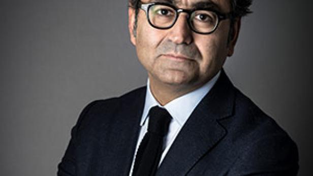 Ein Mann mit Brille und dunklem Anzug vor einem grauen Hintergrund.