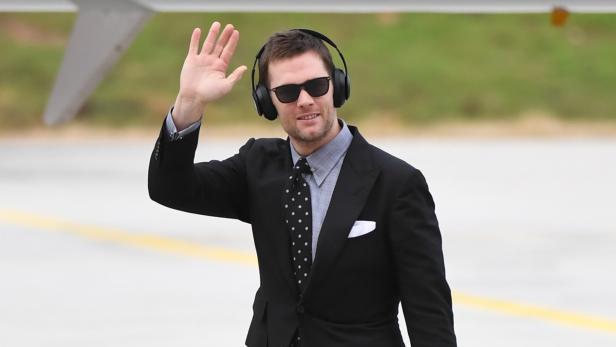 Tom Brady mit Sonnenbrille und Kopfhörern winkt vor einem Flugzeug.