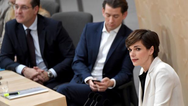 Eine Frau spricht vor Mikrofonen, während zwei Männer im Hintergrund sitzen.