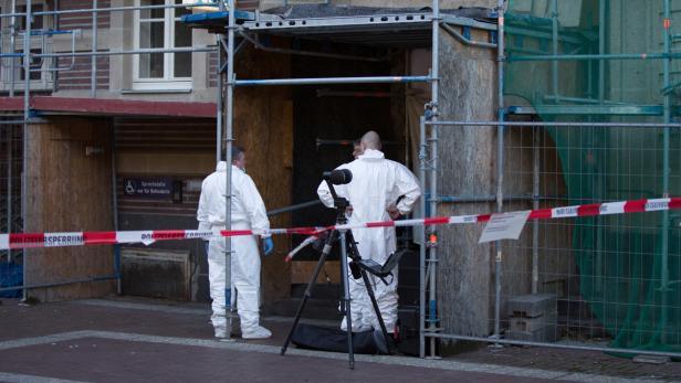 Zwei Personen in Schutzanzügen stehen vor einem Gebäude, abgesperrt mit rot-weißem Band.