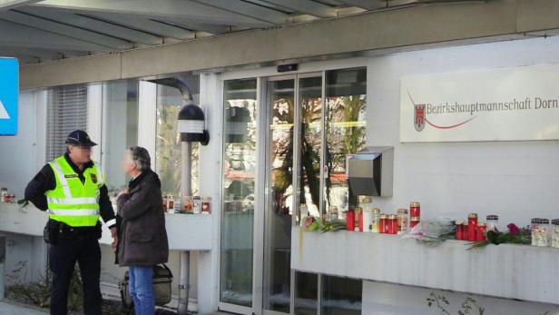 Vor der Bezirkshauptmannschaft Dornbirn stehen ein Polizist und eine Frau neben Kerzen und Blumen.
