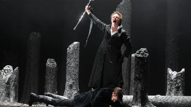 Eine Frau in einem schwarzen Mantel steht mit einem Schwert über einem am Boden liegenden Mann.