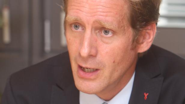 Ein Mann mit blonden Haaren und einem roten Band auf seinem Jackett spricht.