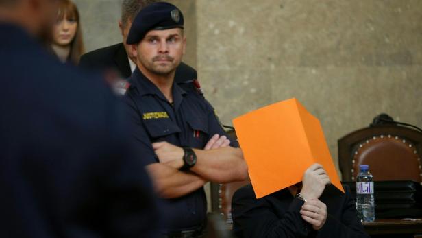 Ein Justizwachebeamter steht neben einer Person, deren Gesicht mit einem orangefarbenen Karton bedeckt ist.