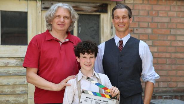 Drei Personen, darunter ein Kind mit Filmklappe, posieren für ein Foto am Set von „Kästner und der kleine Dienstag“.