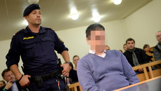 Ein Justizwachebeamter steht neben einer Person mit verpixelten Gesicht in einem Gerichtssaal.