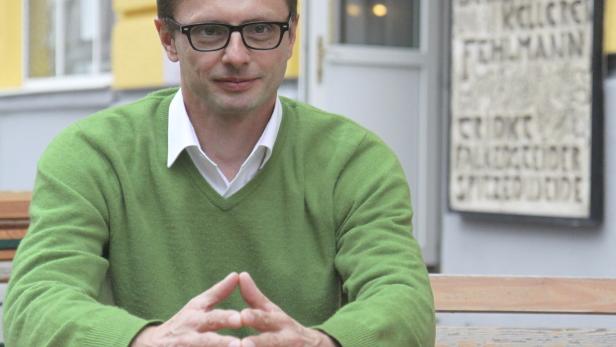 Ein Mann mit Brille und grünem Pullover sitzt an einem Holztisch.