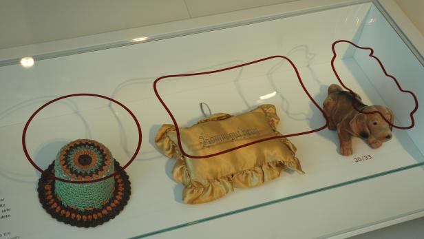 In einer Vitrine liegen eine gehäkelte Dose, ein Kissen mit der Aufschrift „Komm gut heim“ und ein Stofftierhund.