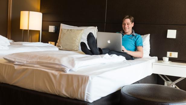 Ein Mann liegt in einem Hotelbett und arbeitet an einem Laptop.