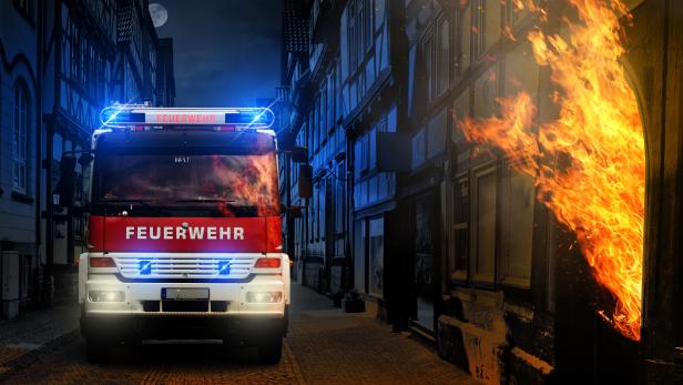 Ein Feuerwehrauto mit Blaulicht steht vor einem brennenden Gebäude.