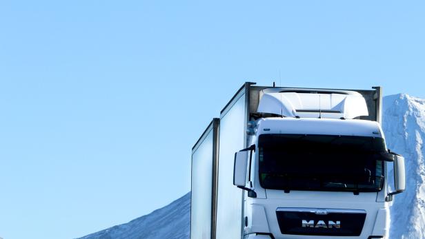 Ein weißer MAN-Lkw fährt auf einer asphaltierten Straße vor schneebedeckten Bergen.