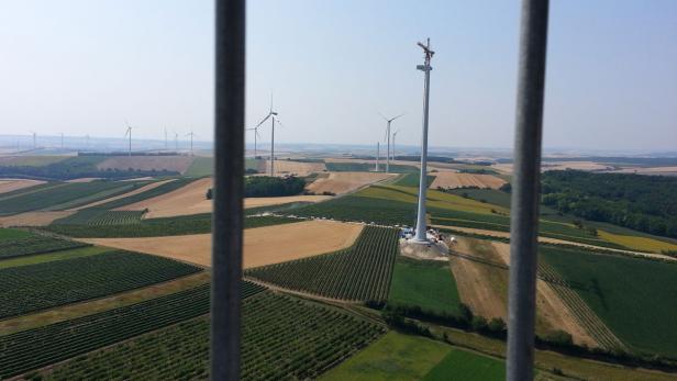 Blick durch Gitterstäbe auf eine Landschaft mit Windrädern, von denen eines im Bau ist.