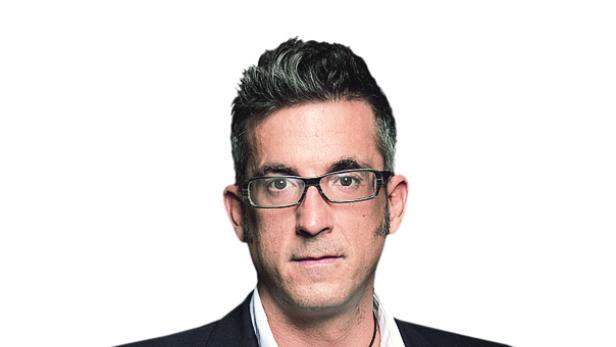 Ein Mann mit Brille, Sakko und Jeans posiert mit den Händen in den Hosentaschen.