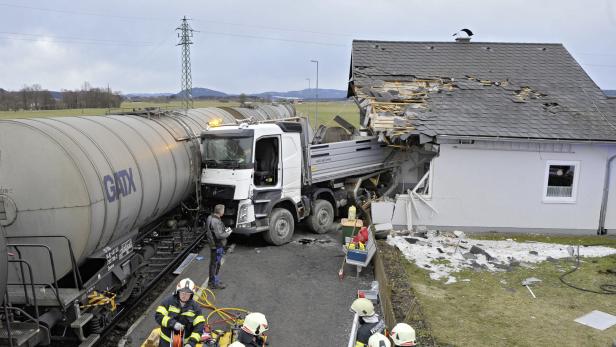 Ein Lastwagen ist in ein Haus gefahren, während ein Zug daneben steht; Feuerwehrleute sind vor Ort.
