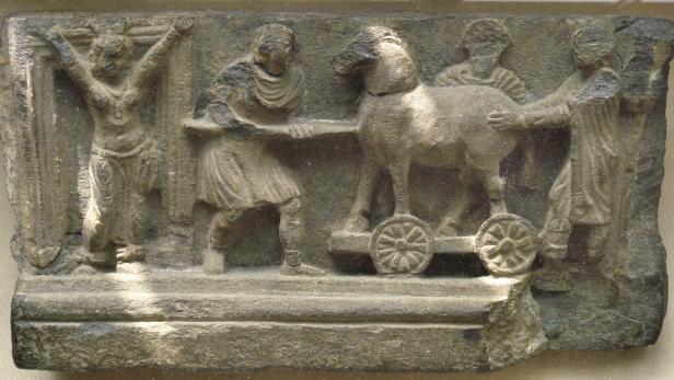 Ein Fries mit der Darstellung eines hölzernen Pferdes auf Rädern, das von mehreren Figuren bewegt wird.