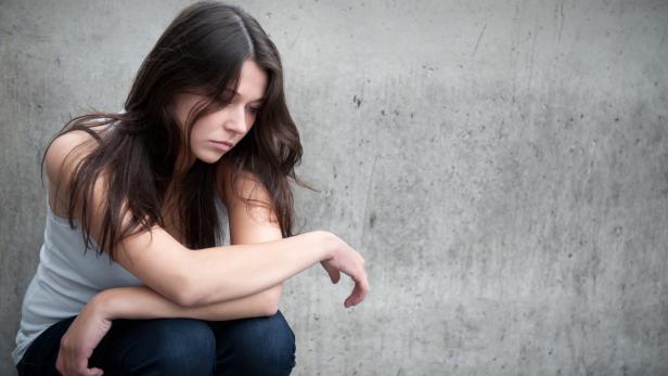 Eine junge Frau sitzt niedergeschlagen vor einer grauen Wand.