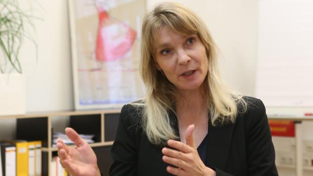 Eine blonde Frau in einem schwarzen Blazer gestikuliert während eines Gesprächs.